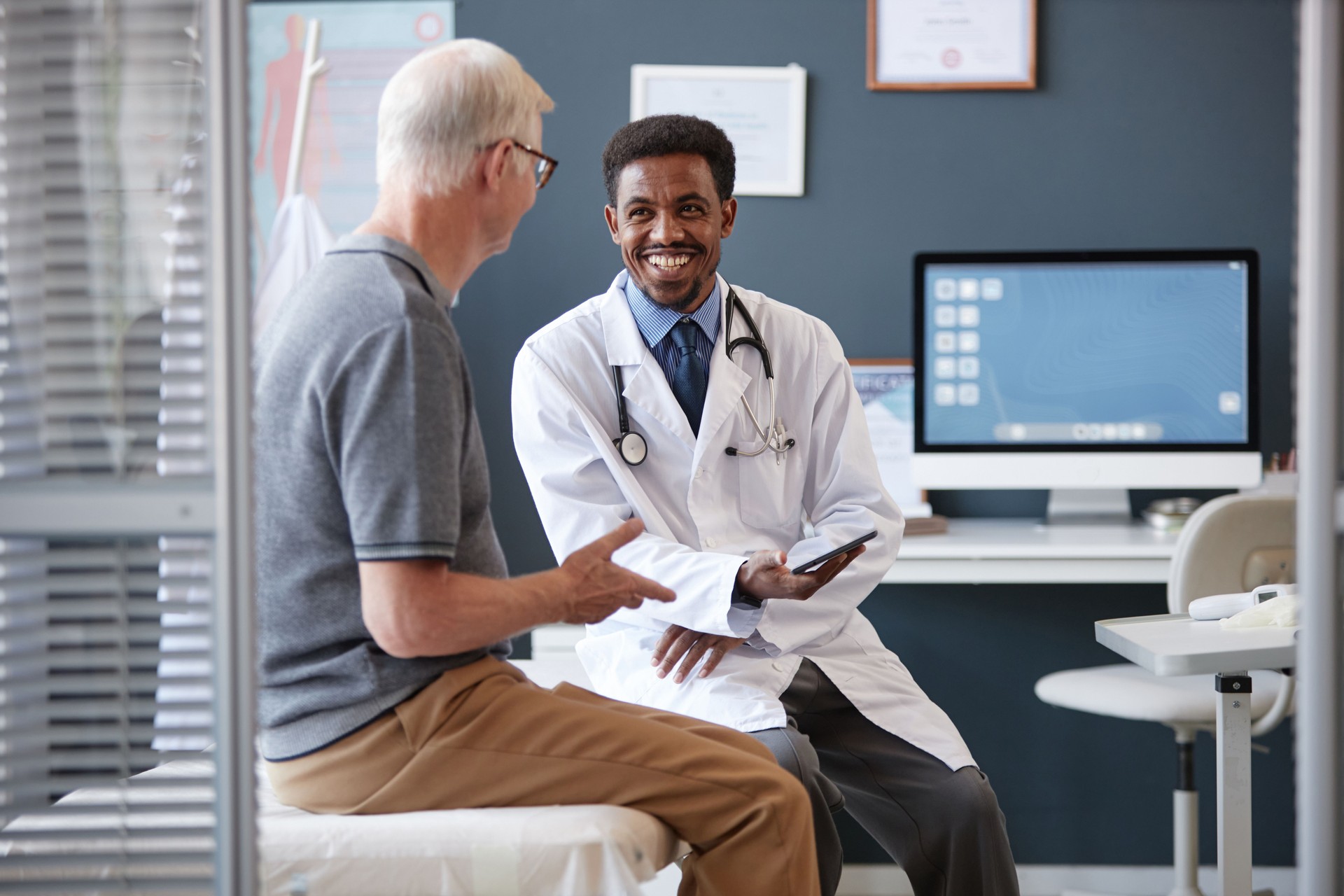 Smiling Black Doctor Holding Tablet Talking to Senior Patient in Consultation